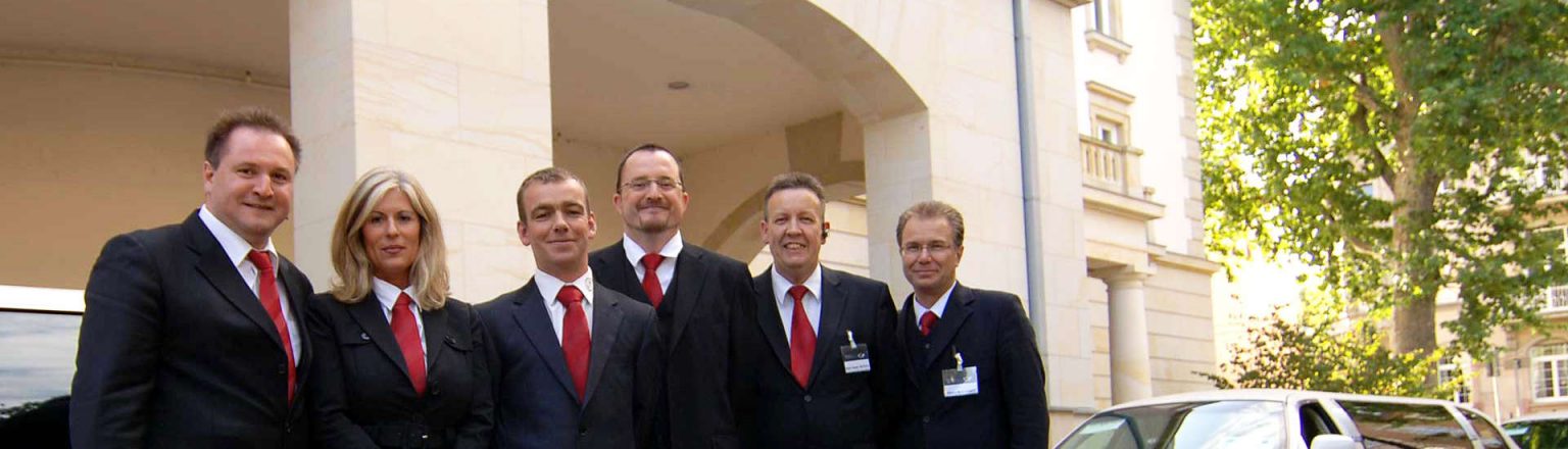 Fünf Männer und eine Frau in dunklen Anzügen mit roten Krawatten von der Firma German Limousines.