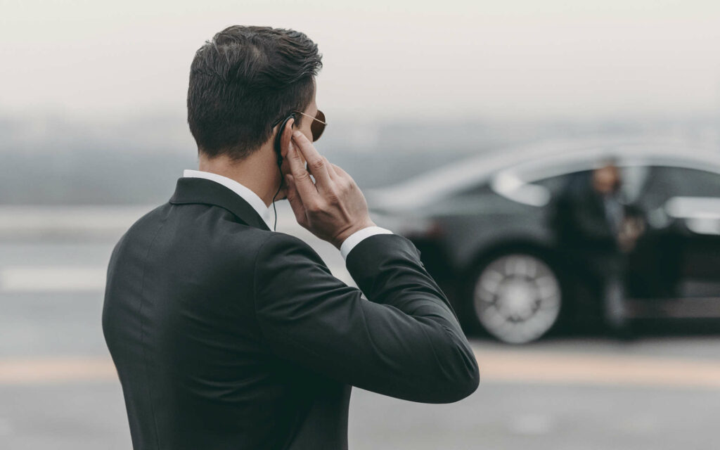 Ein Sicherheits Mann, der mit einem Anzug bekleidet ist, hört mit einem Headset jemandem, zu und beobachtet einen Mann, der aus einem schwarzen Auto aussteigt.