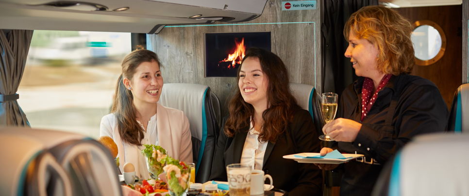 bistro-bus-bordkueche Zei junge Frauen werden im Lounge Liner von German Limousines Network GmbH & Co. KG mit Getränken und Essen versorgt. So wird ein Messebesuch mit der ganzen Firma zu einem Incentive.